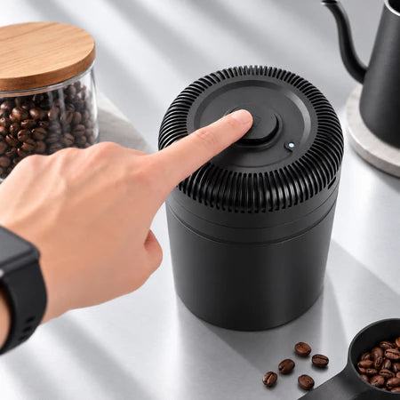 A black cylindrical coffee grinder with a ribbed top and a finger pressing the central button, shown on a light countertop with coffee beans and a kettle nearby.